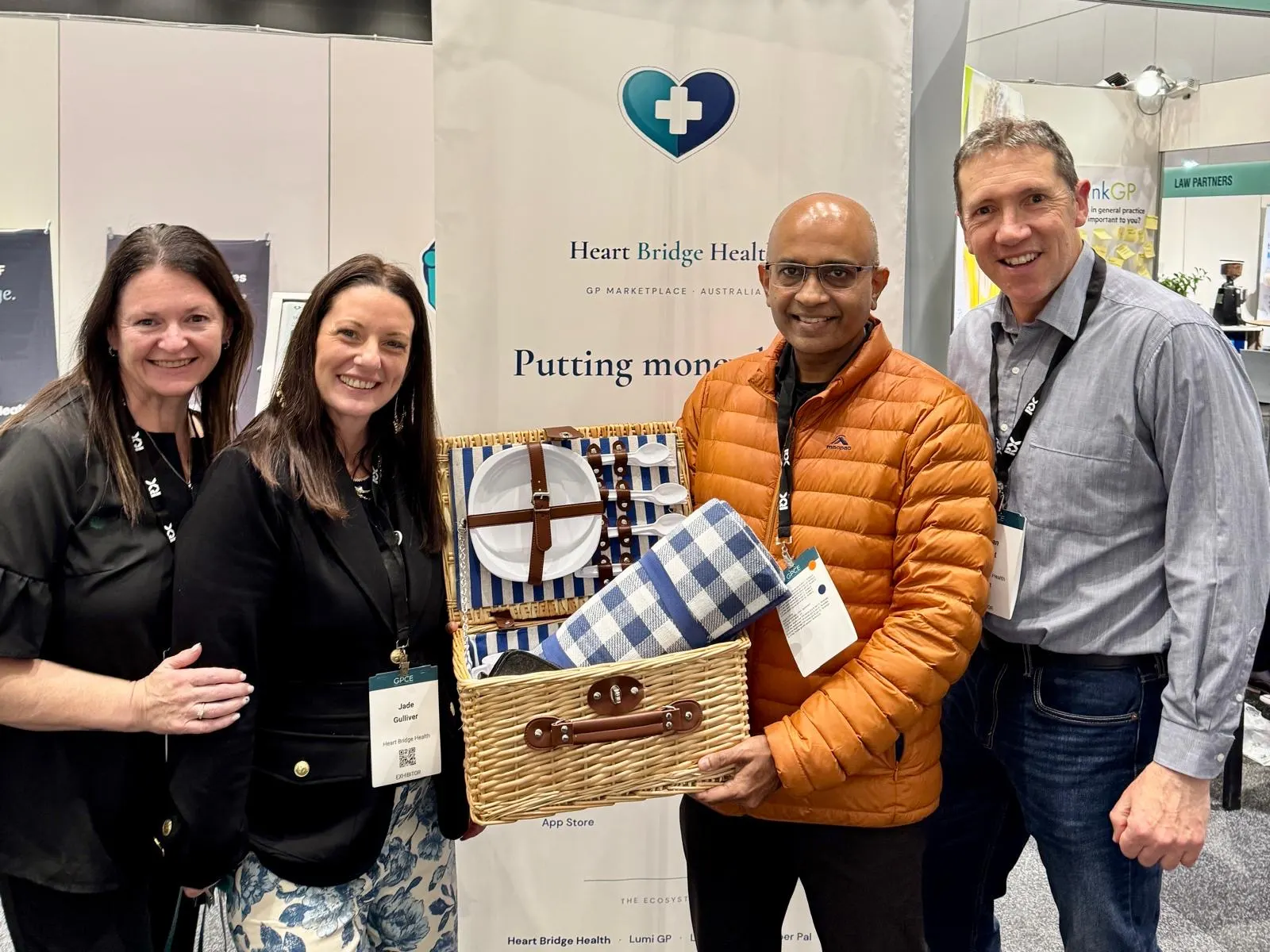 Four people smiling together at the Heart Bridge Health conference stand, holding a picnic hamper prize