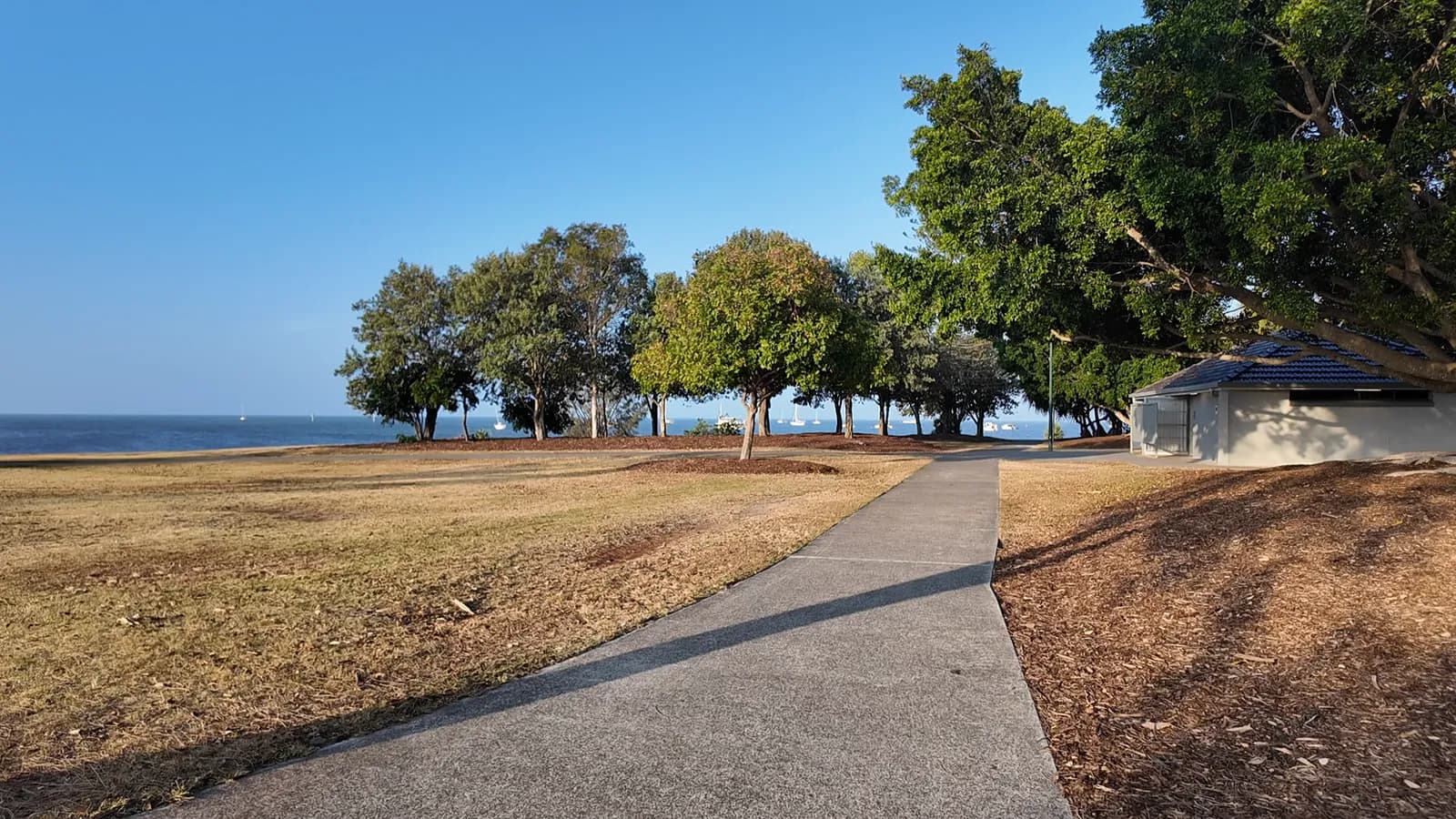 A sunny foreshore park in Cleveland with a footpath, shady trees and the calm water of Moreton Bay