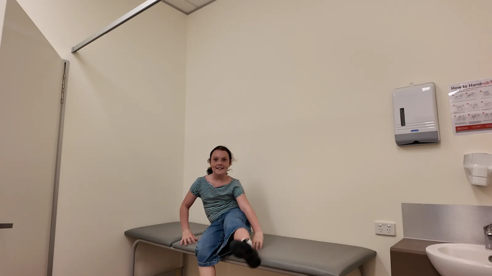 A clean, bright consulting room with an examination couch, a sink and a paper towel dispenser, and a girl sitting on the couch