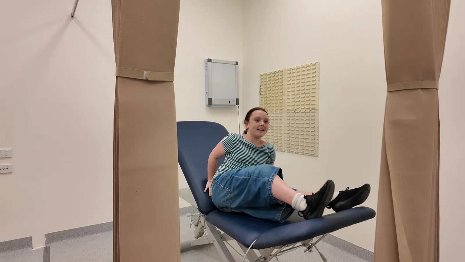 A treatment room with a blue examination chair, curtains and a wall-mounted dressing station, with a girl sitting on the chair and smiling
