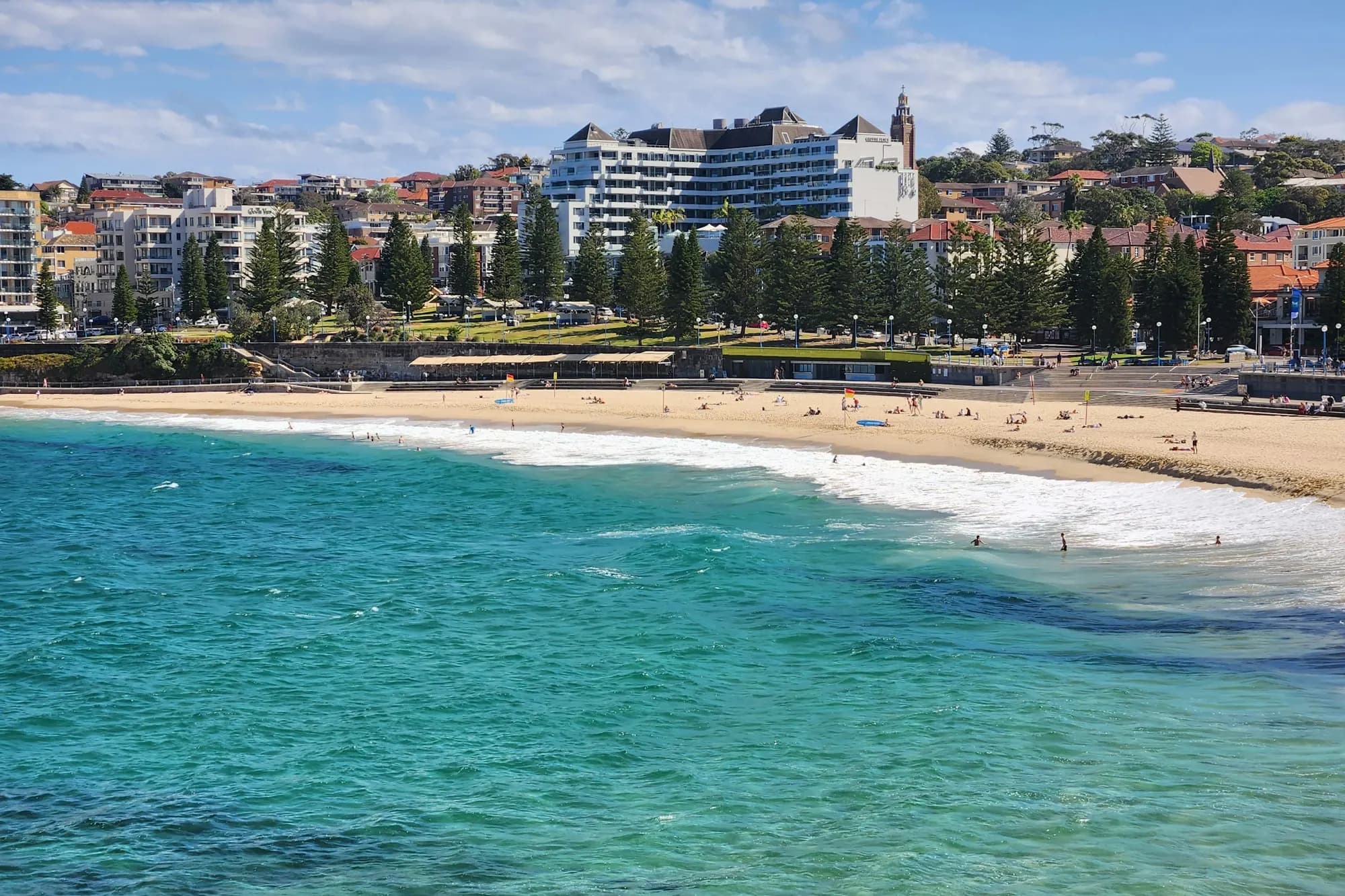 A wide view of Coogee Beach, turquoise water breaking onto the sand with the promenade, Goldstein Reserve and the suburb rising behind it