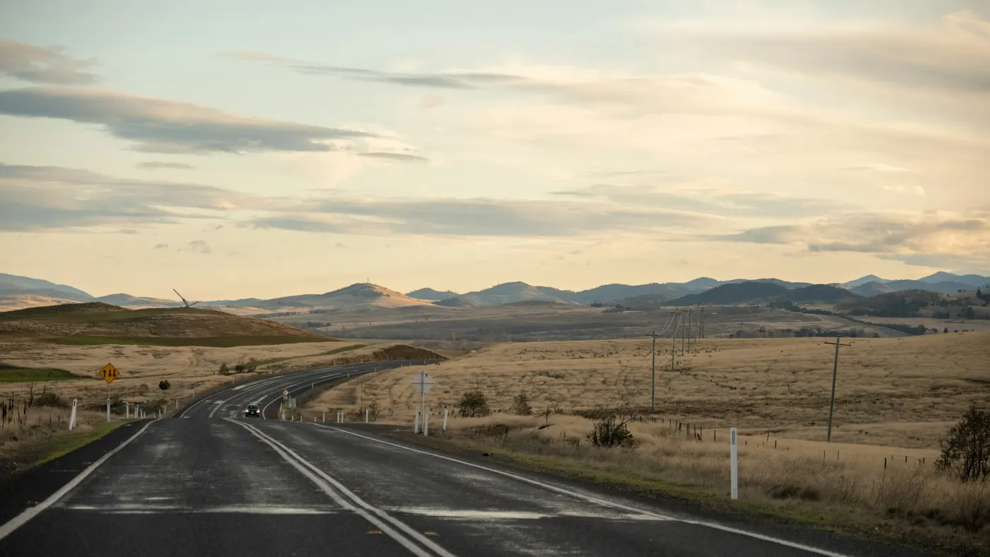 An open country road running through dry Australian farmland towards low hills on the horizon
