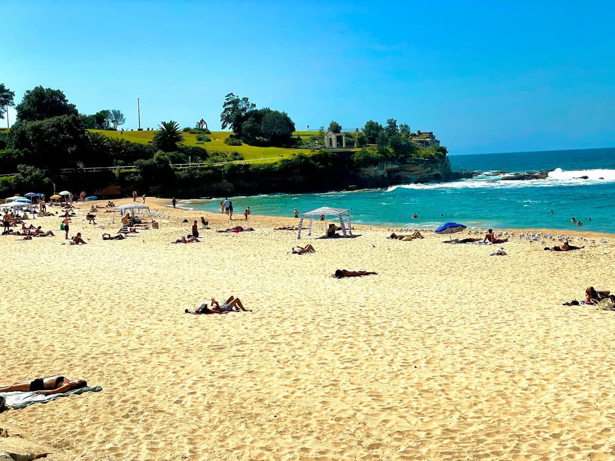 People on the sand at Coogee Beach, with the grassy headland and the old baths at the northern end behind them