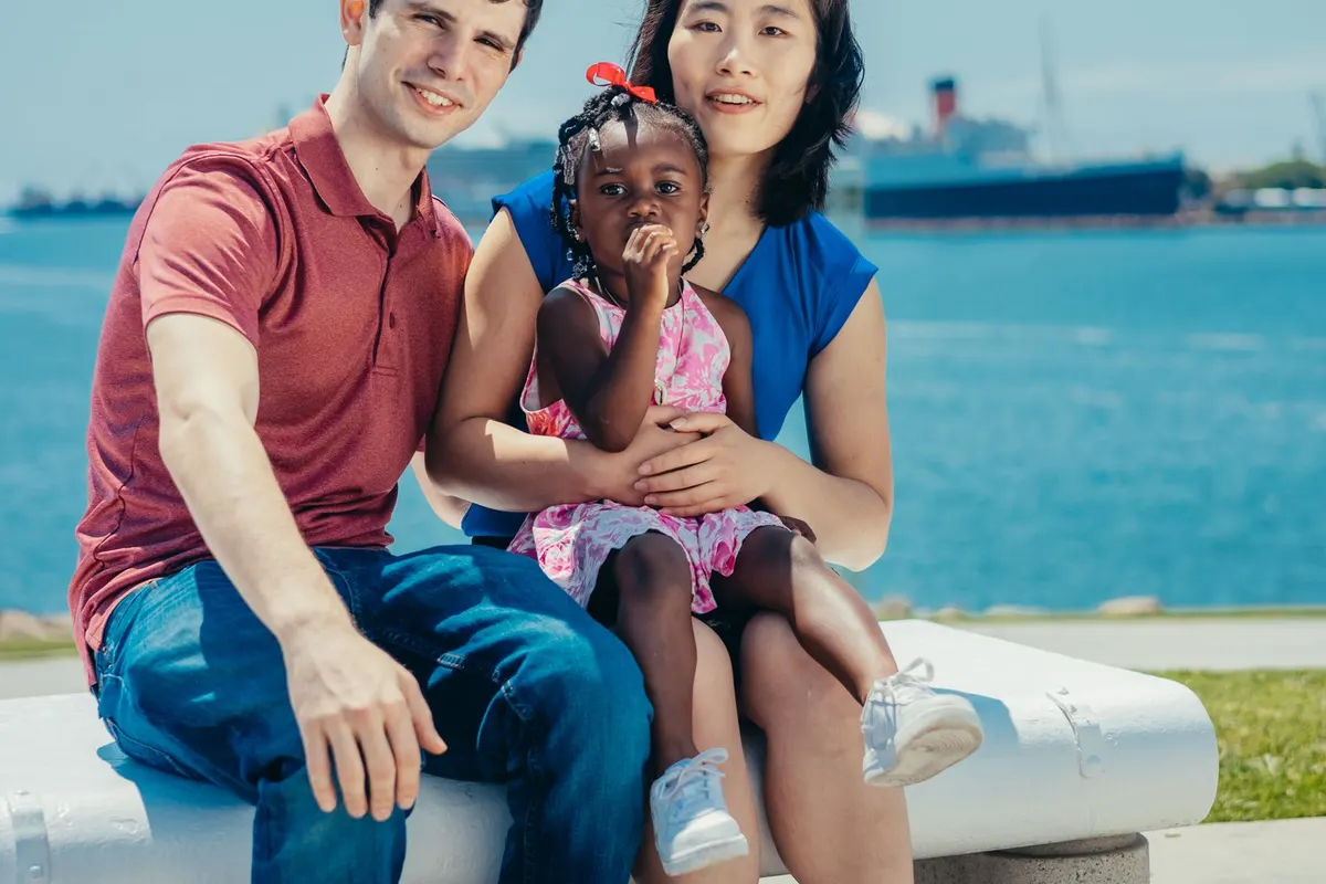 A family of three sitting together by the water on a sunny day, the parents and their young daughter laughing