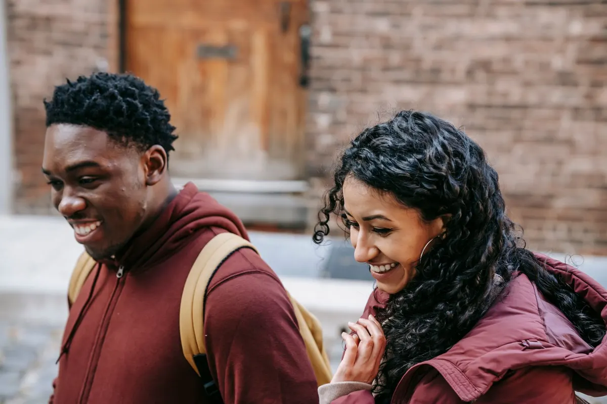 Two people standing close together outdoors, one leaning in and smiling as the other laughs
