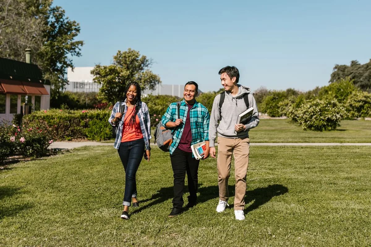 Three university students walking together across a sunny campus lawn, carrying books and backpacks