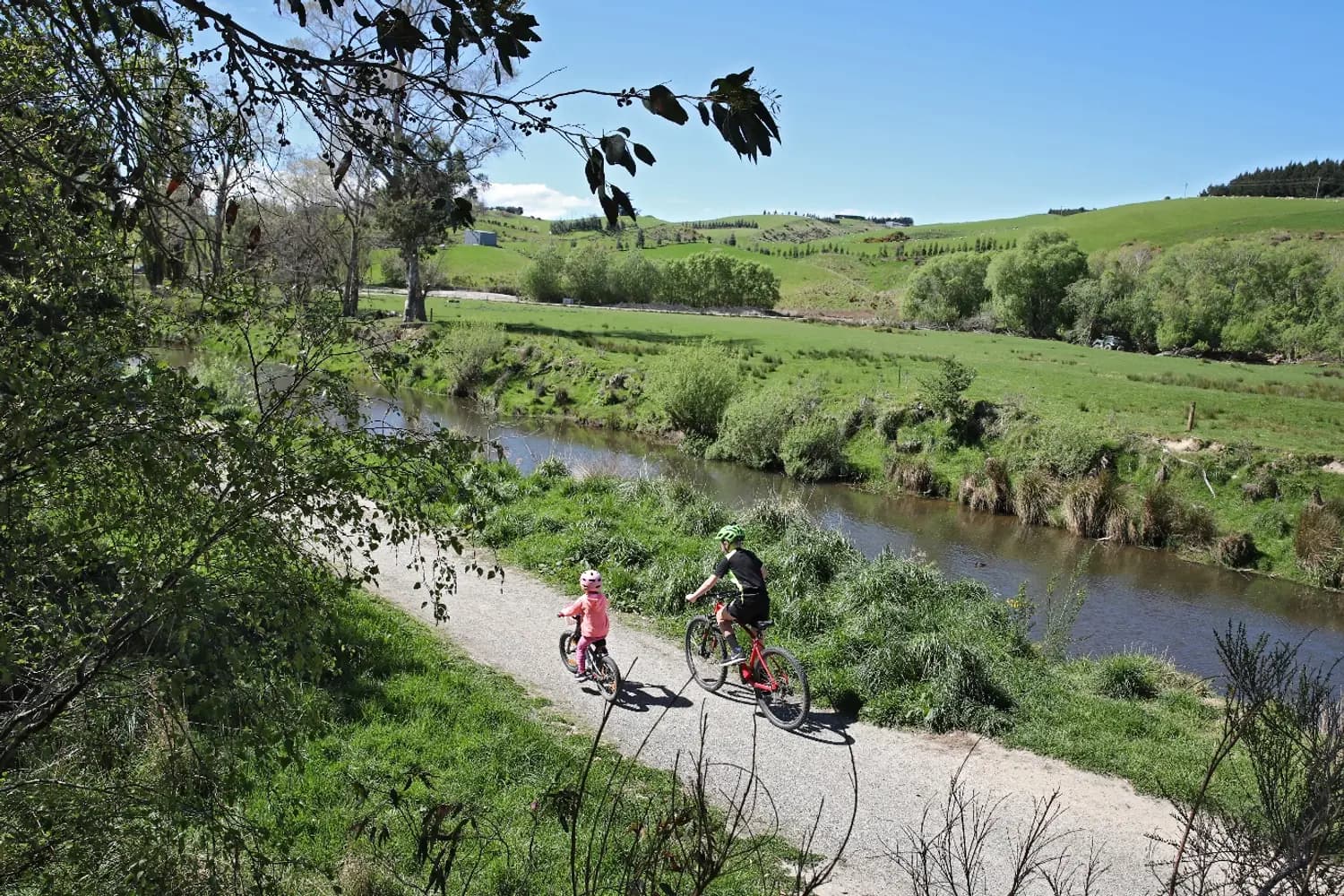 A parent and small child riding bikes on a gravel river trail through green Southland farmland